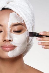 Young asian half-naked lady with towel on head put scrab on her face with brush isolated over grey